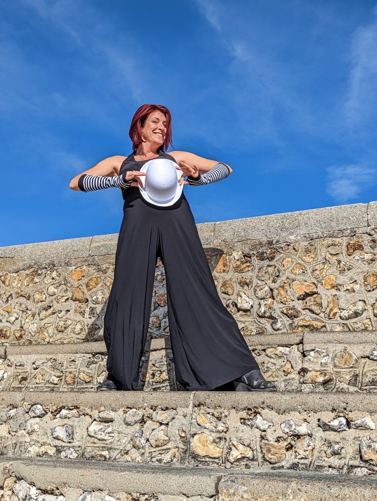 Lex in black trouser suit wearing stripped fingerless gloves and holding a white bowler hat to her chest.